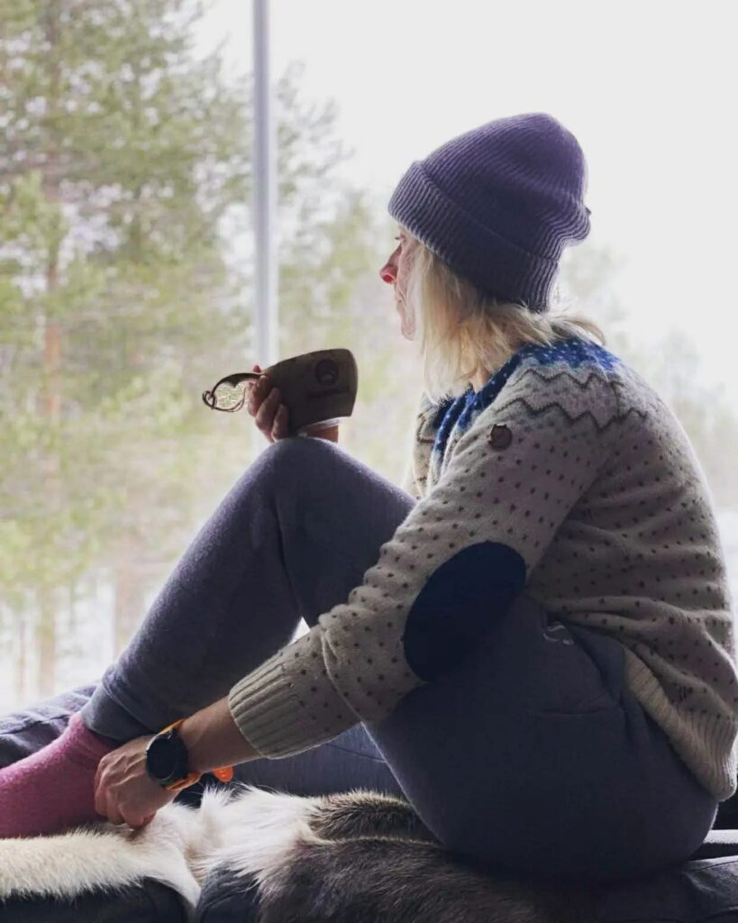 Dea Larsen sidder ved et vindue med en kop i hånden og ser ud over naturen under refleksionerne før deltagelsen i Alene i Vildmarken.