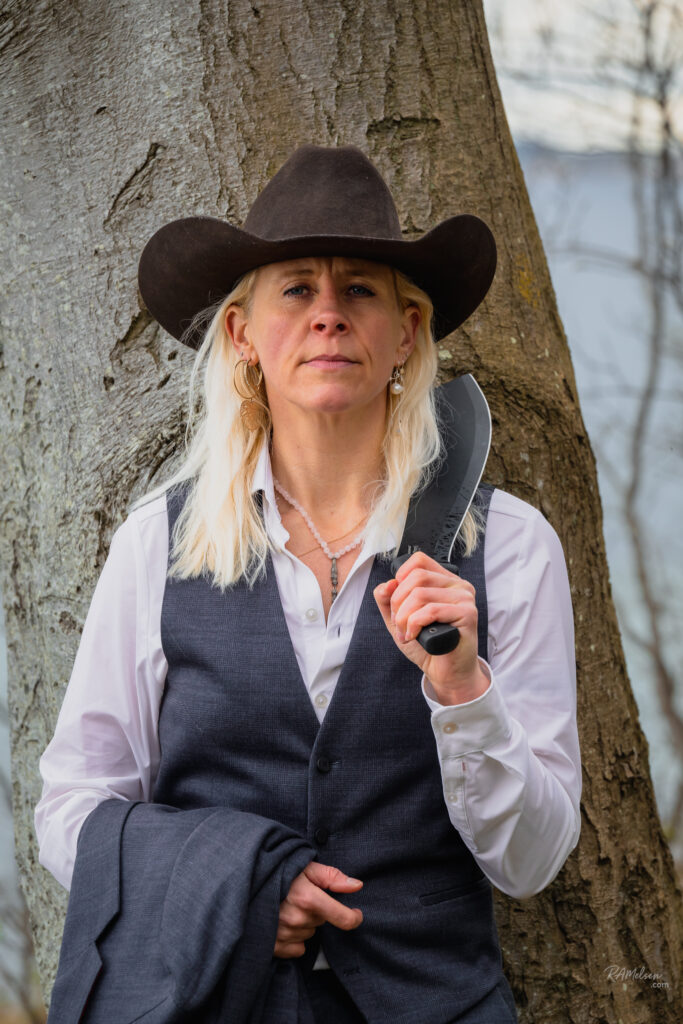 Portræt af Dea Larsen med cowboyhat og kniv ved et træ. Kendt fra Alene i Vildmarken sæson 10 og passioneret friluftsmenneske.