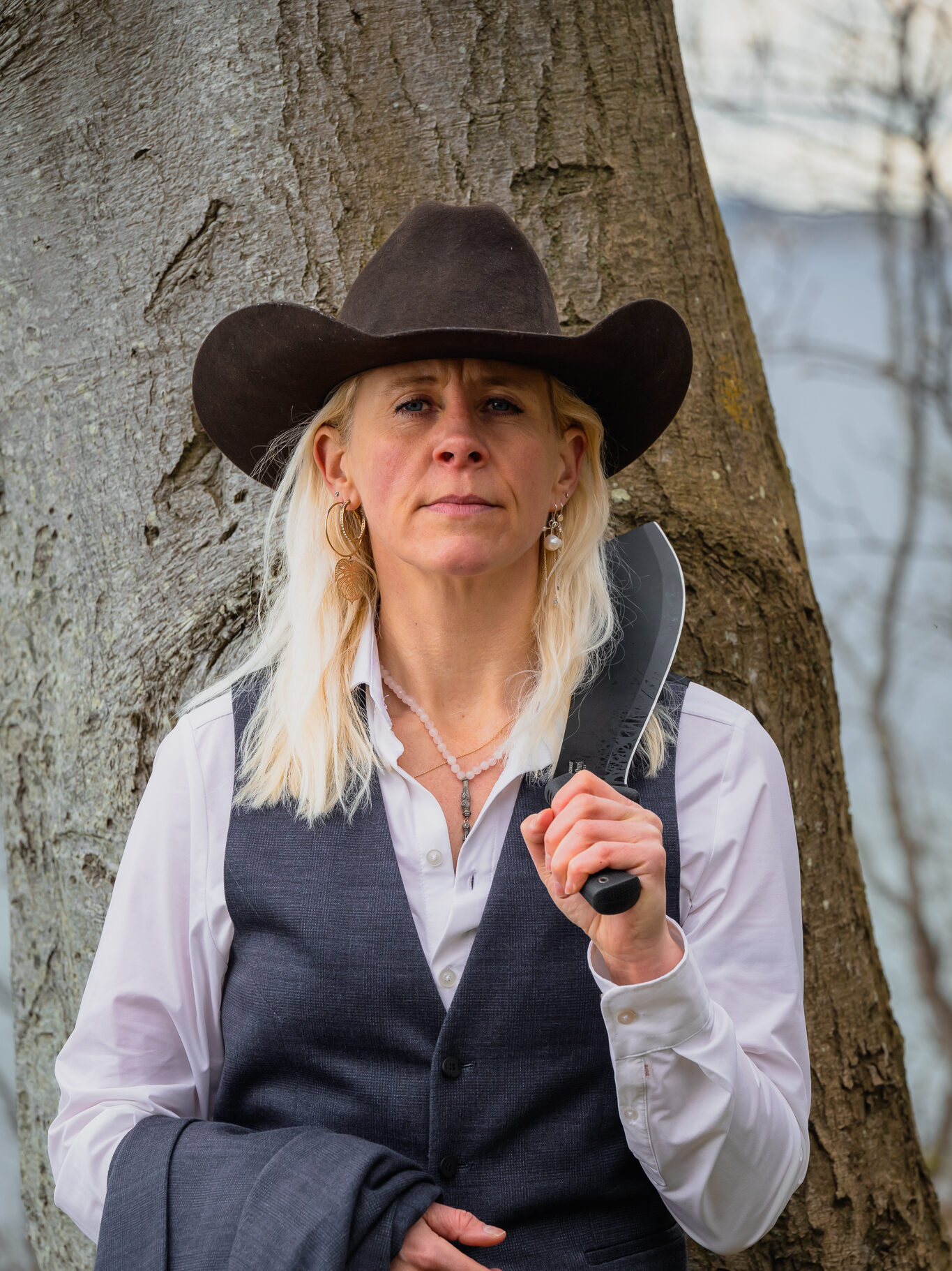 Portræt af Dea Larsen med cowboyhat og kniv ved et træ. Kendt fra Alene i Vildmarken sæson 10 og passioneret friluftsmenneske.