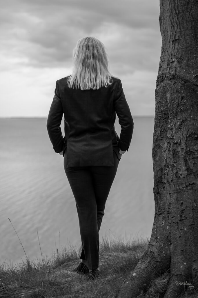 Sort-hvidt portræt af Dea Larsen stående ved vandet med ryggen til og blikket mod horisonten. Billedet symboliserer ro, eftertanke, friluftsliv og nye eventyr efter Alene i Vildmarken.