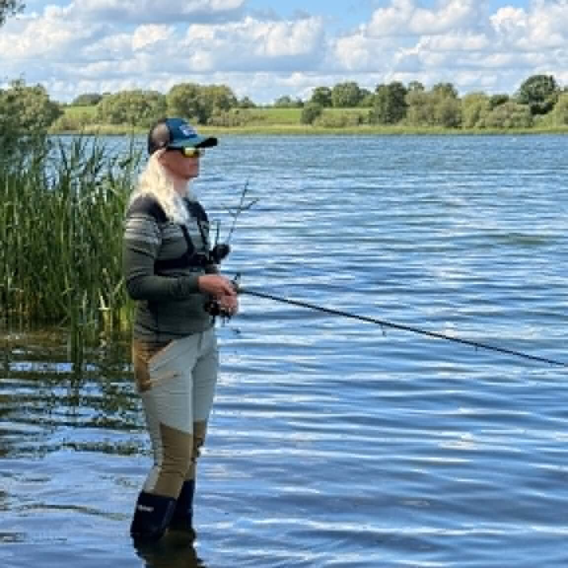 Dea Larsen står i vandkanten og fisker på en stille sommerdag omgivet af siv, blå himmel og rolige omgivelser.
