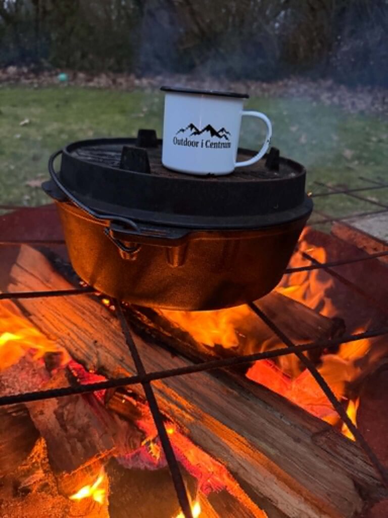 Bål med glødende brænde, Dutch Oven og en hvid emaljekop fra Outdoor i Centrum. Billedet symboliserer friluftsliv, nærvær og de enkle glæder ved livet under åben himmel
