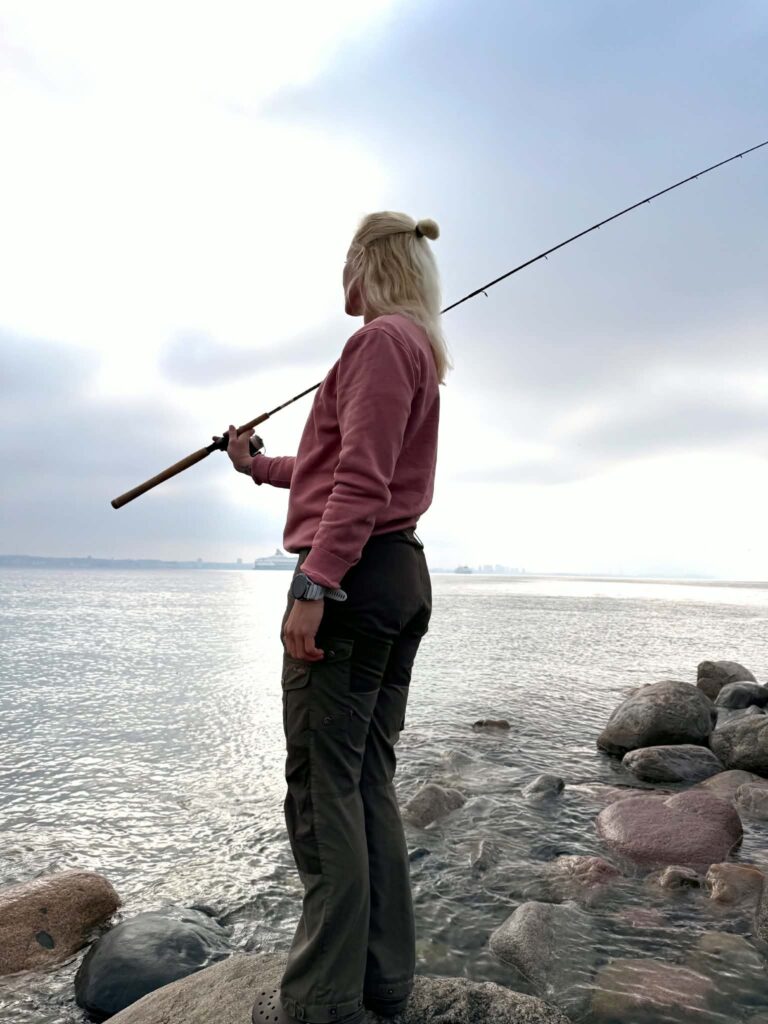 Dea Larsen står ved vandet med en fiskestang og nyder roen. Billedet symboliserer de erfaringer og livslektioner, hun tog med sig hjem fra Alene i Vildmarken