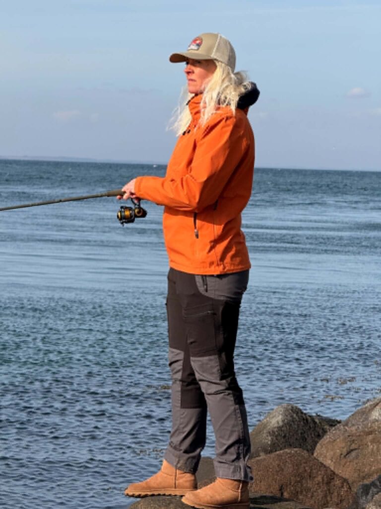 Dea Larsen i orange jakke ved havet. Kendt fra Alene i Vildmarken sæson 10, foredragsholder, lystfisker og indehaver af Outdoor i Centrum.